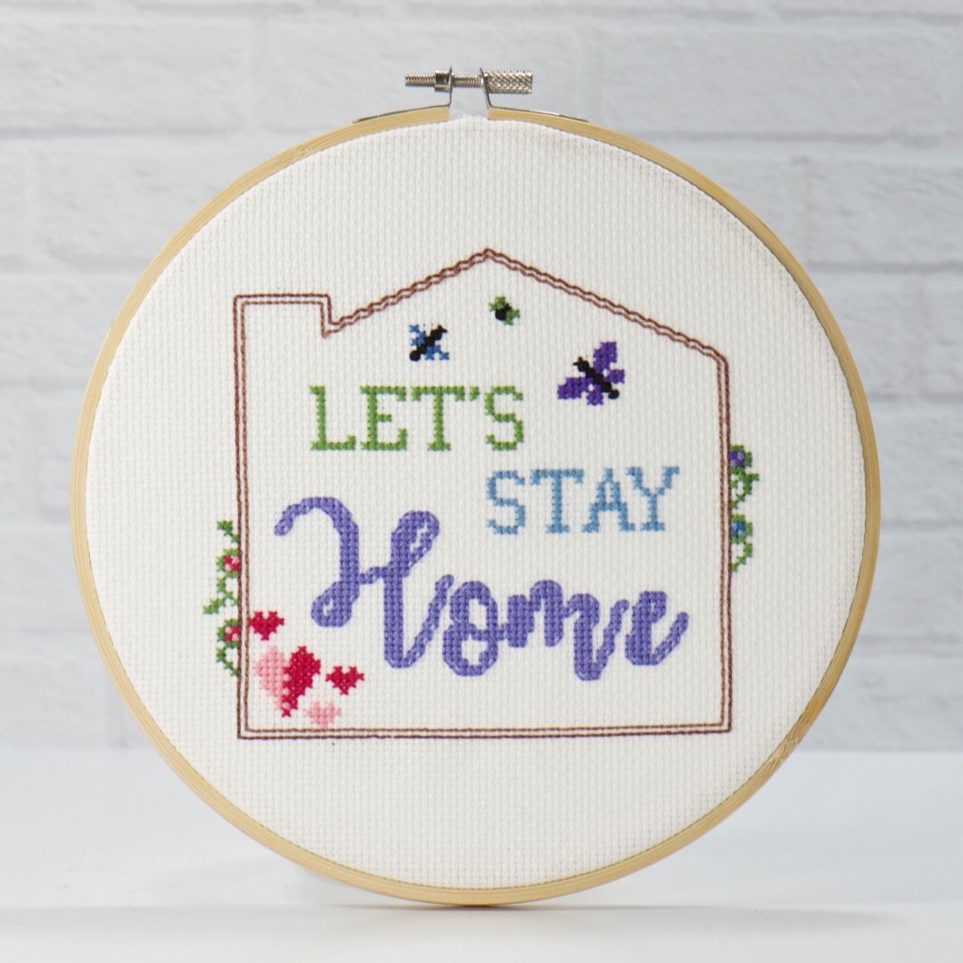 Let's Stay Home words surrounded by blue and purple butterflies, pink and red hearts.  Featuring a back stitched outline with vines peeking around the outside of this cross stitch pattern
