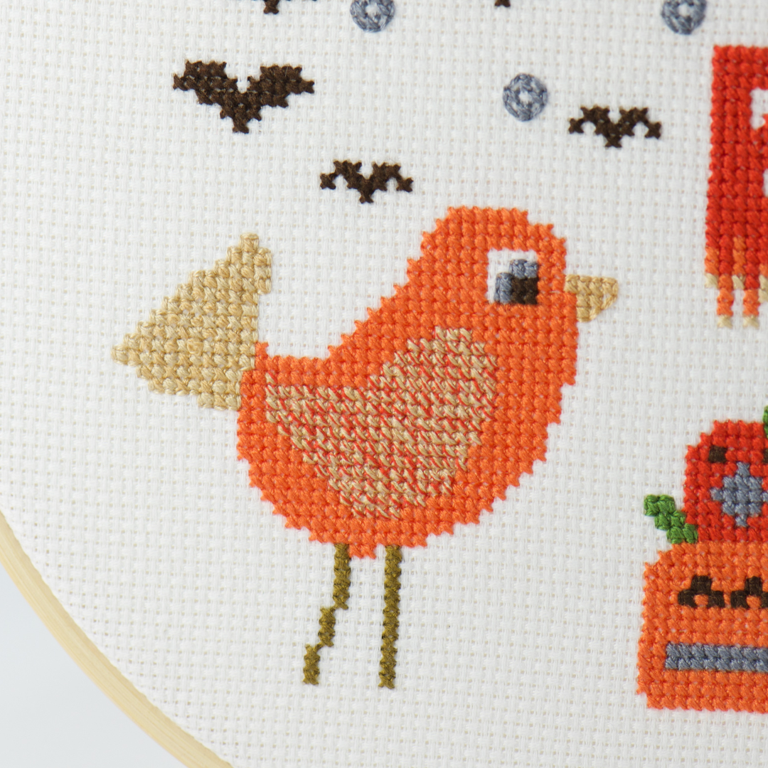 cross stitch bird with multi colored wing inquisitively looking at three orange pumpkins