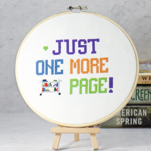cross stitch words Just One More Page with a colorful book cart on white cross stitch fabric inside a wood embroidery hoop